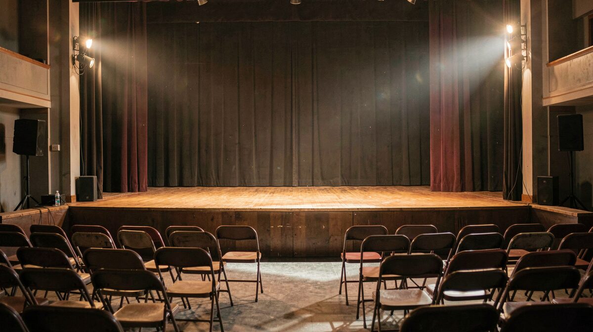 Un escenario de teatro comunitario vacío con sillas plegables en primer plano, luz cálida de focos laterales, encuadre desde el patio de butacas.